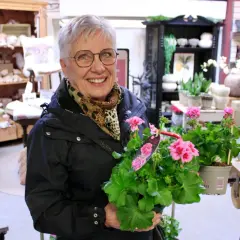 Glad kvinna med årets pelargon i famnen, omgiven av blommor och inredningsdetaljer i en butik.