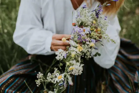 En person i vit blus och randig kjol håller en bukett med sju sorters blommor, en midsommartradition.