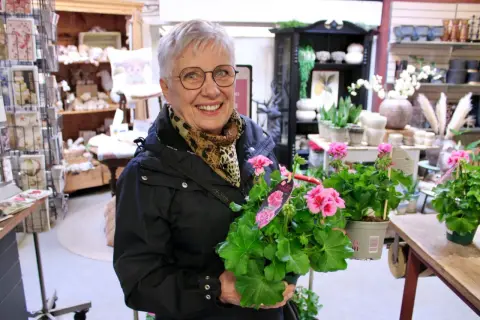 Glad kvinna med årets pelargon i famnen, omgiven av blommor och inredningsdetaljer i en butik.