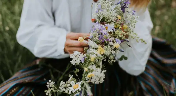 En person i vit blus och randig kjol håller en bukett med sju sorters blommor, en midsommartradition.