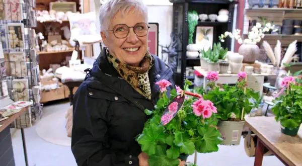 Glad kvinna med årets pelargon i famnen, omgiven av blommor och inredningsdetaljer i en butik.