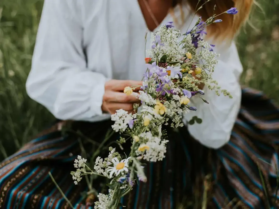 En person i vit blus och randig kjol håller en bukett med sju sorters blommor, en midsommartradition.