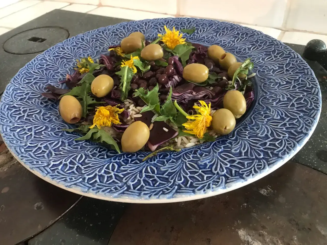 En bl&aring;m&ouml;nstrad tallrik med en sallad av ris, svarta b&ouml;nor, r&ouml;dk&aring;l, oliver och maskrosblommor. Maskros nyttigt!